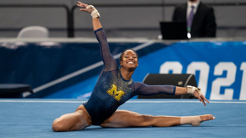 Sierra Brooks, a dark-skinned Black woman wearing a dark blue leotard with a large yellow letter M on the chest, poses in a bent-knee split position with her right arm over head while smiling widely.