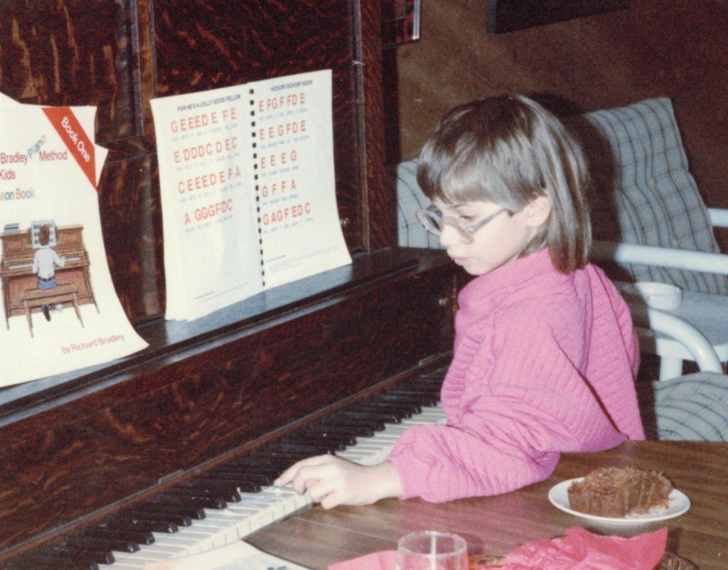 A fair-skinned child with shoulder-length brown hair and thick glasses, wearing a pink quilted jacket, plays an upright piano while resting their arm on a table behind them.